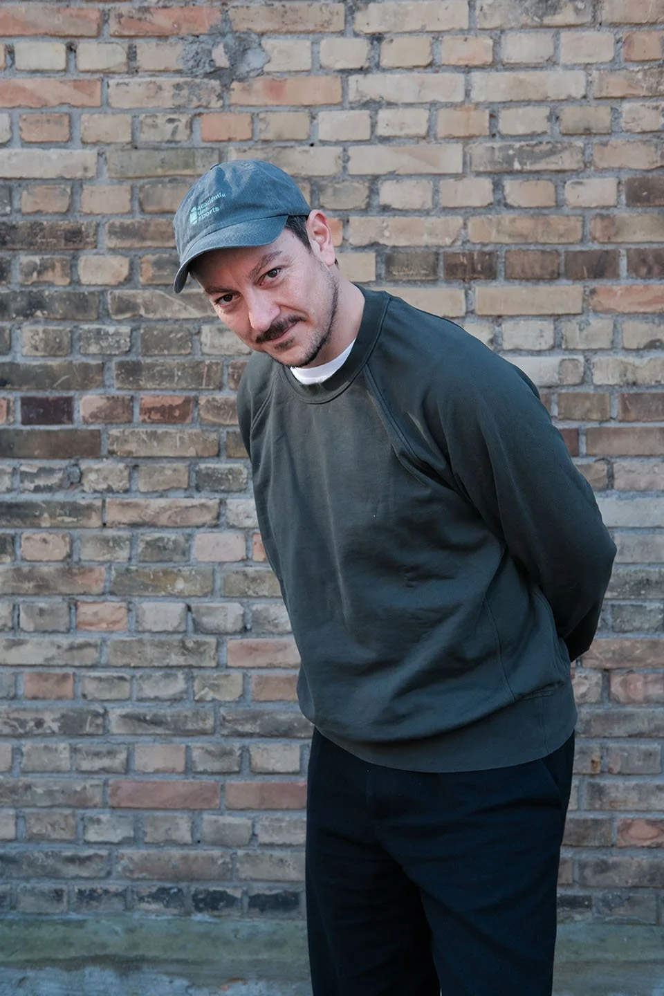 Ein Mann steht vor einer Backsteinmauer, trägt eine dunkle Jacke und einen dunkelgrauen Baseballcap, sieht in die Kamera und hat die Hände im Rücken verschränkt.