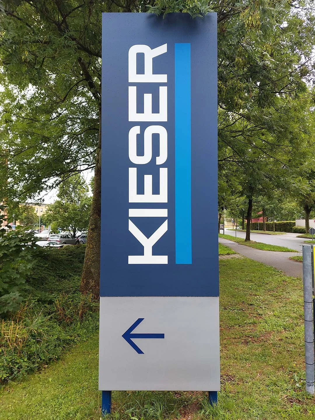 Ein Werbepylon mit der Aufschrift 'Kiesser' und einem Pfeil nach links, umgeben von Bäumen und einem Bürgersteig.