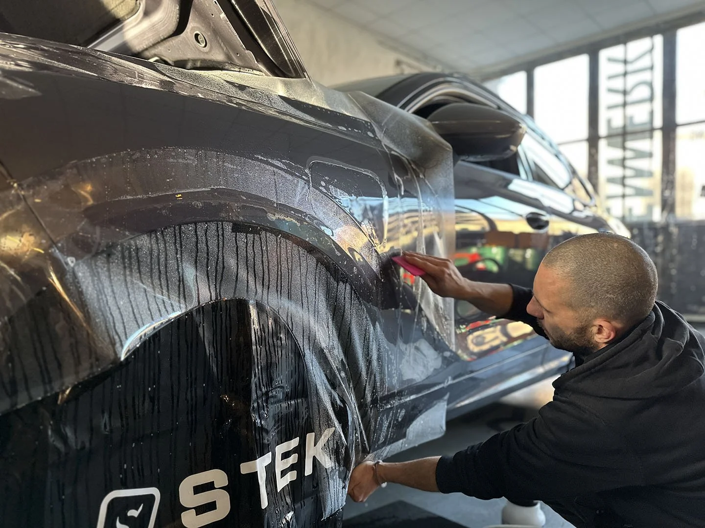 Ein Mann klebt mit einem Werkzeug einen Aufkleber auf die Seite eines schwarzen Autos.