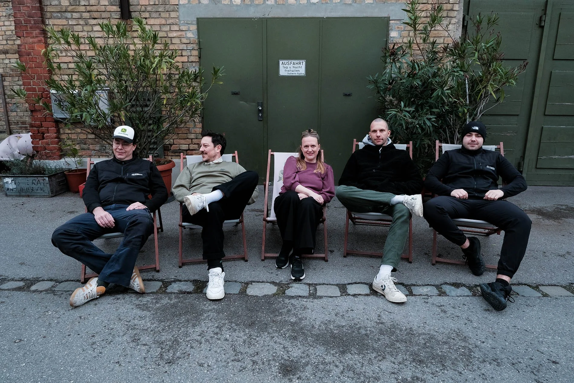 Fünf Menschen sitzen in Liegestühlen vor einer grünen Tür, umgeben von Pflanzen und einer Ziegelwand.
