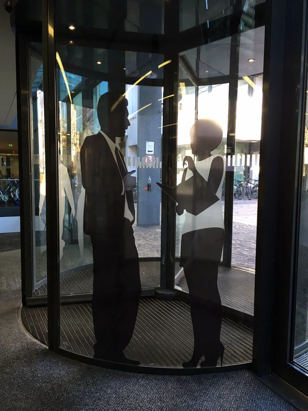 Silhouetten von zwei Personen, einem Mann und einer Frau, die in einem Eingangsbereich eines Gebäudes miteinander sprechen. Der Mann trägt einen Anzug, die Frau trägt ein Kleid und hohe Schuhe.
