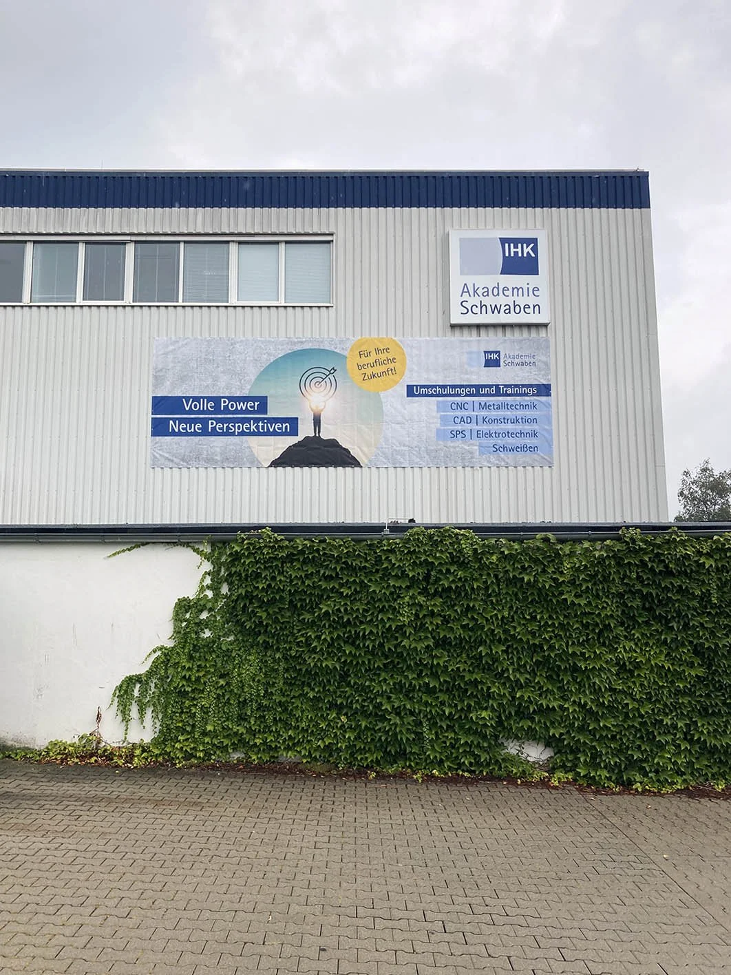 Das Bild zeigt die Außenansicht eines Gebäudes mit einem großen Banner, das die IHK Akademie Schwaben bewirbt, inklusive eines Slogans und einer Liste von Kursen. Es gibt Pflanzen vor der Wand und ein Fenster im oberen Bereich.
