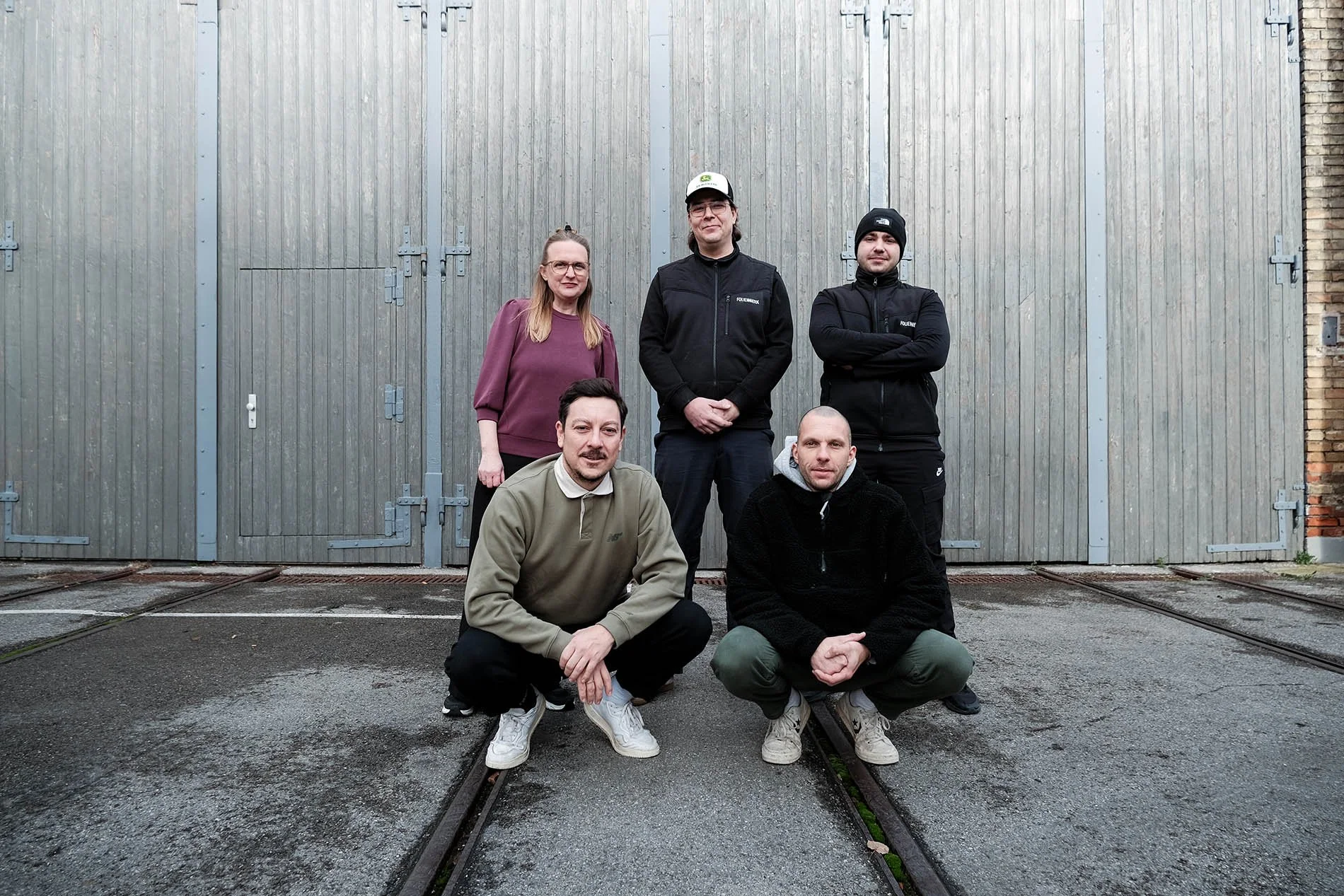 Das Folienwerk-Team: Fünf Menschen vor einer großen grauen Tür, drei stehen und zwei knien. Alle schauen in die Kamera.