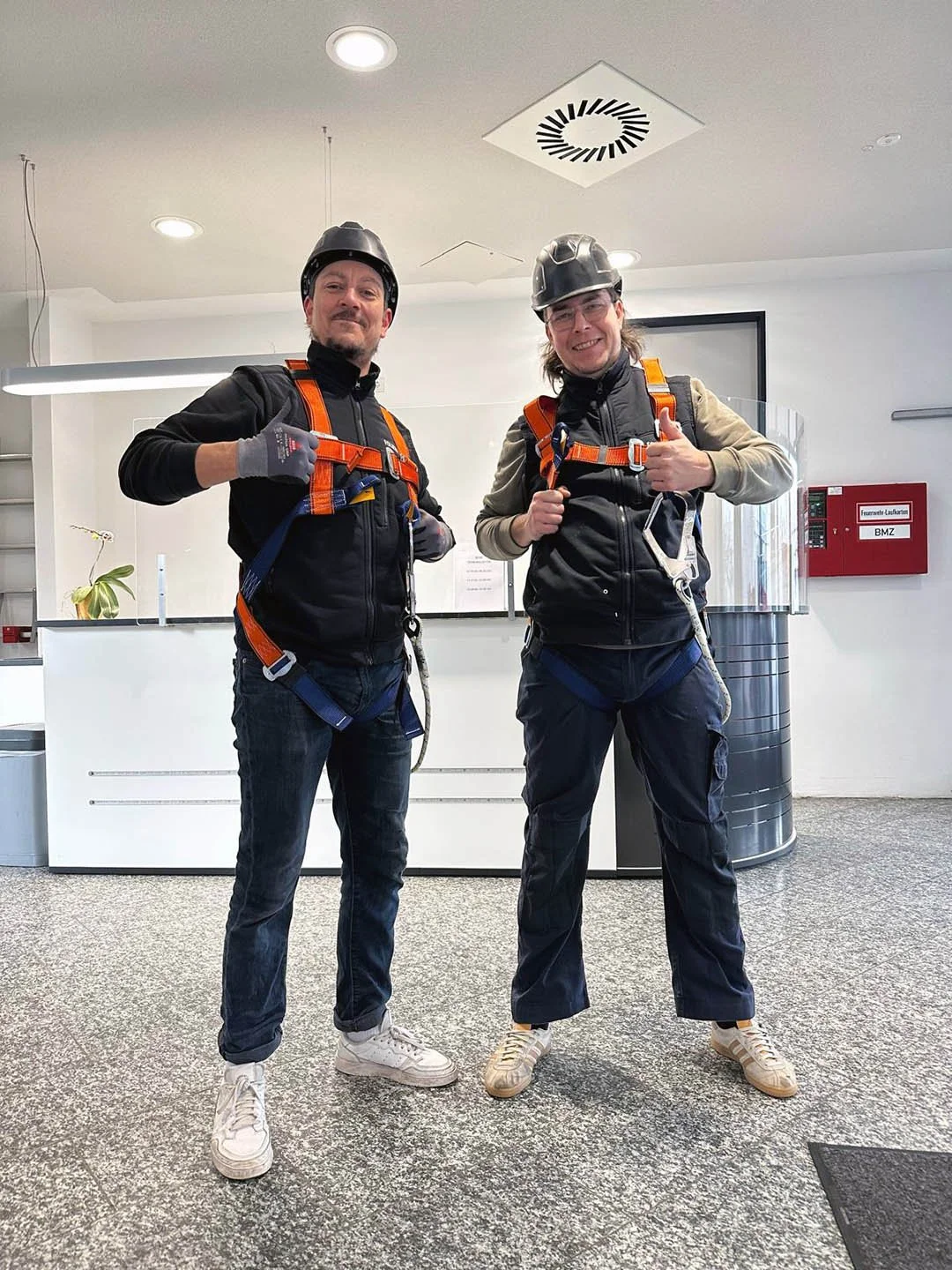 Zwei Männer in Schutzhelmen und Sicherheitsgurten stehen im Büro, beide lächeln und zeigen positiv. Einer trägt einen schwarzen Anzug, der andere einen beige Jacke, beide tragen Sneakers.