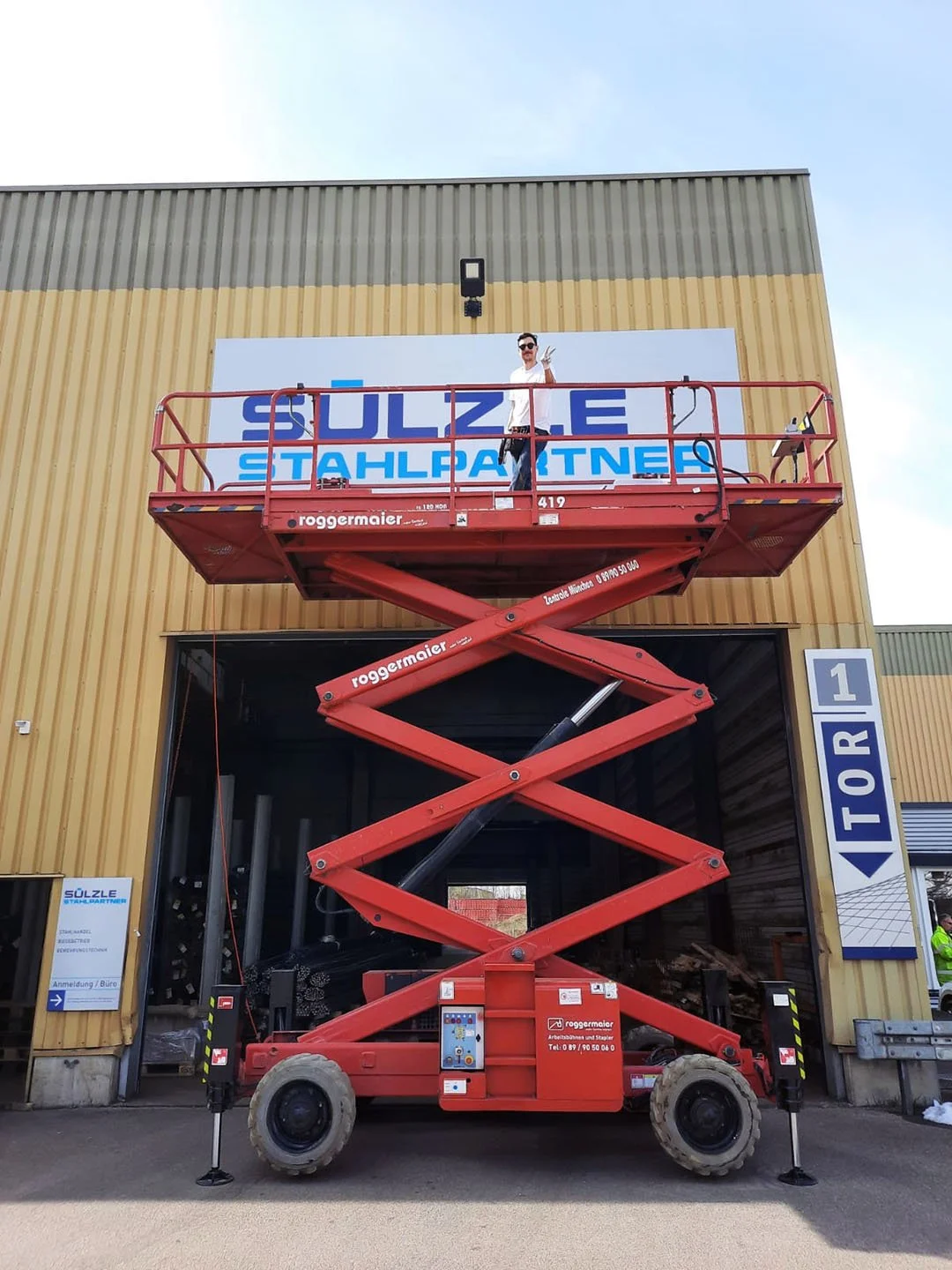 Eine Person steht auf einem roten Hebebühnenwagen vor einem Gebäude mit einem großen Schild. Das Gebäude hat eine gelbe und grüne Fassade. Der Himmel ist teilweise sichtbar und klar.