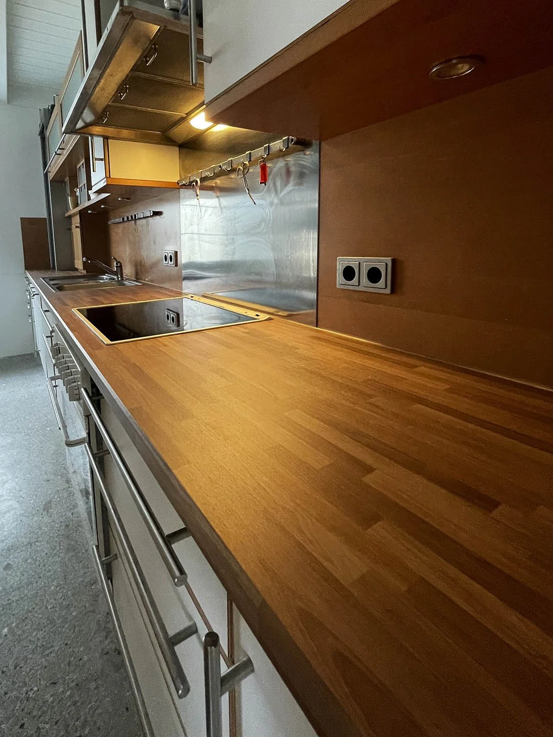 Küchenarbeitsplatte aus Holz mit Herd und Stromanschlüssen, Backofen und Küchenschrank, kleine Küchenutensilien hängen an einer Stange, helle Wände und hoher Deckenbereich.