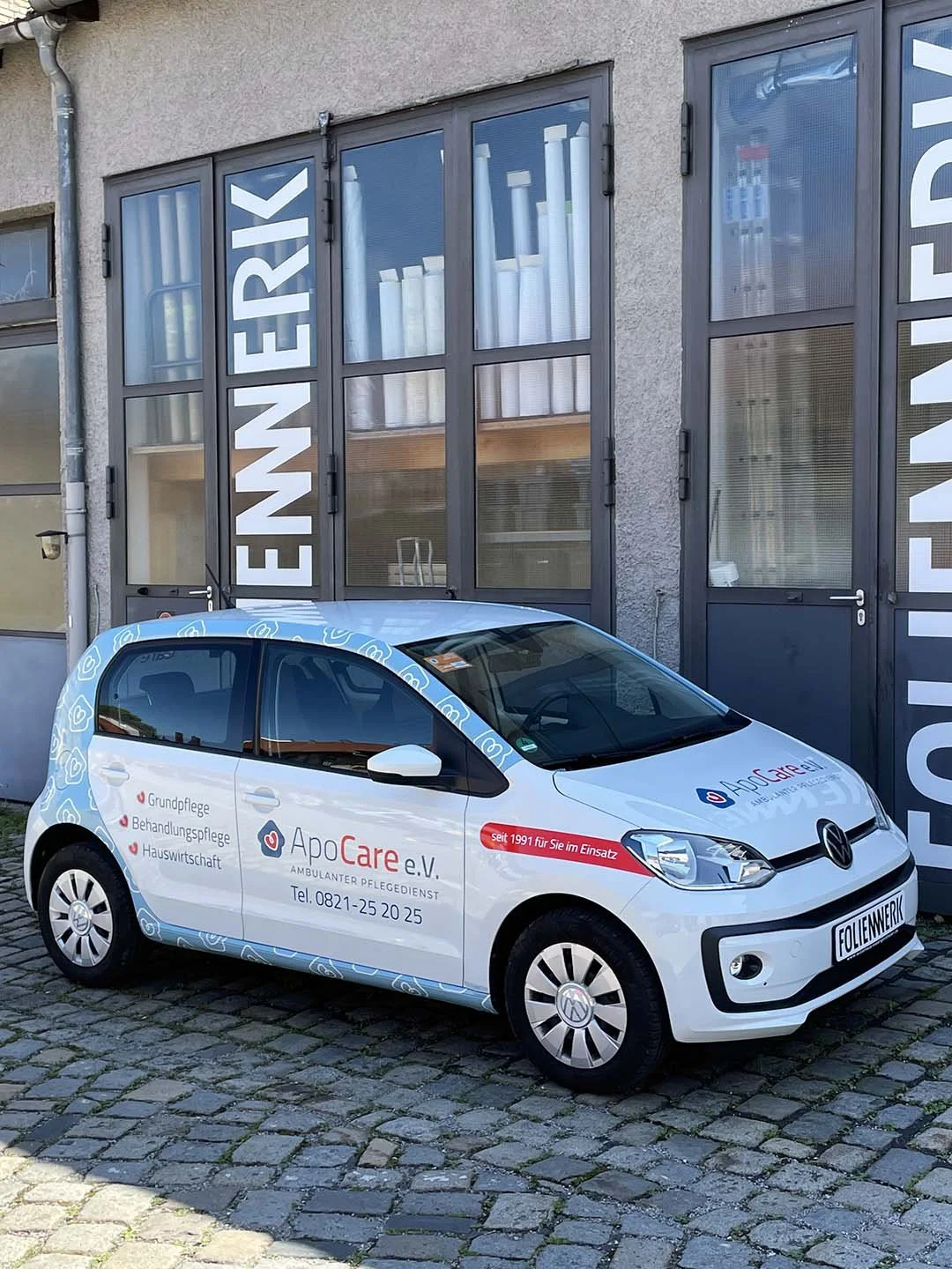 Ein weißer VW Polo mit Beschriftung der Firma ApoCare e.V. ist vor einem Gebäude mit großen Fenstern
