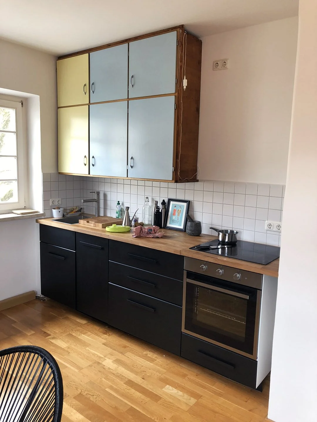 Moderner Küchenraum mit schwarzen Unterschränken, Holzarbeitsplatte, weißen Wandfliesen und bunten Hängeschränken in Hellblau und Gelb