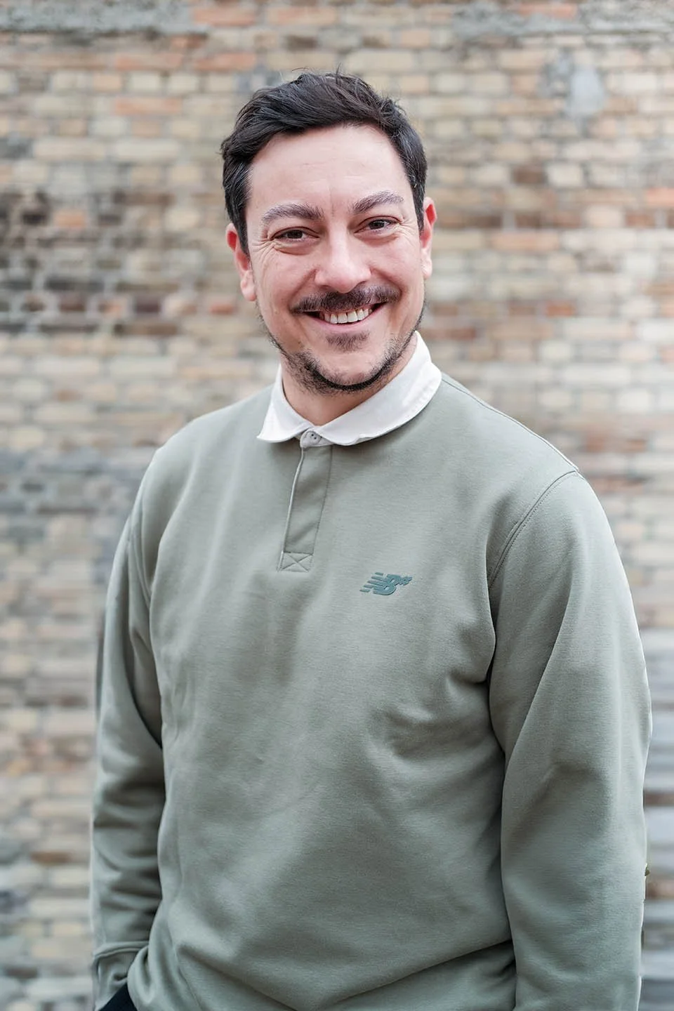 Lächelnder Mann in hellgrüner New Balance Pullover vor einem Ziegelwand-Hintergrund.