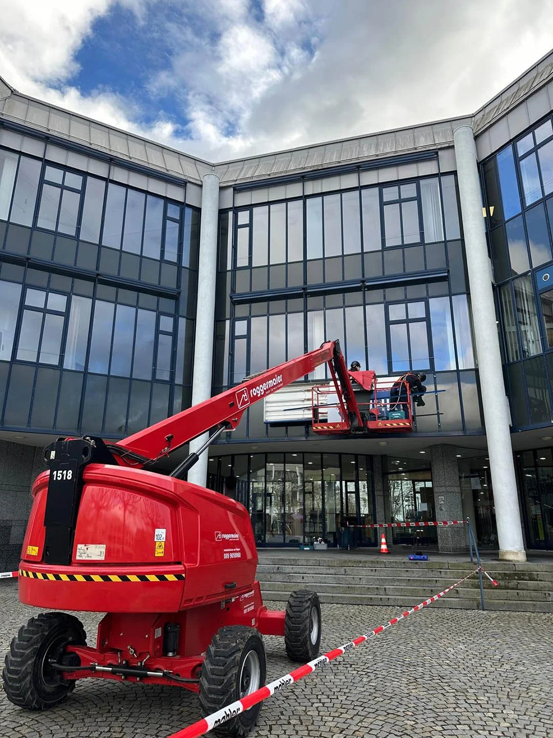 Arbeit mit einem roten Kran auf einem modernen Glasgebäude im Freien, eine Person arbeitet hoch oben an der Fassade, vor dem Gebäude sind Absperrband und Verkehrshütchen platziert.