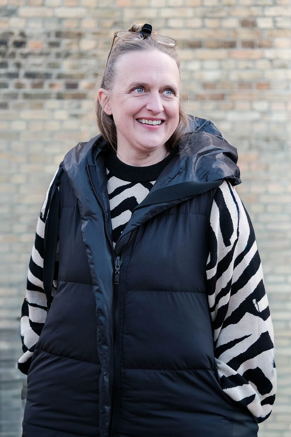 Eine lachende Frau mit blauen Augen, braunem Haar und Brille auf dem Kopf, trägt einen schwarzen, gepolsterten Weste und ein Zebra-Muster-Sweatshirt, vor einer Backsteinmauer.