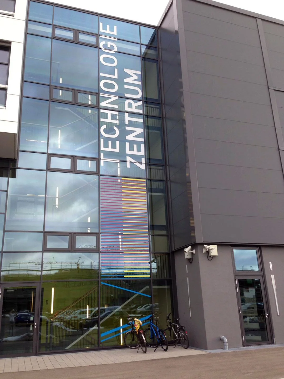 Außenansicht eines modernen Gebäudes mit großen Glasfenstern. An der Fassade steht in weißen Buchstaben 'TECHNOLOGIE ZENTRUM'. Vor dem Gebäude sind zwei Fahrräder abgestellt.