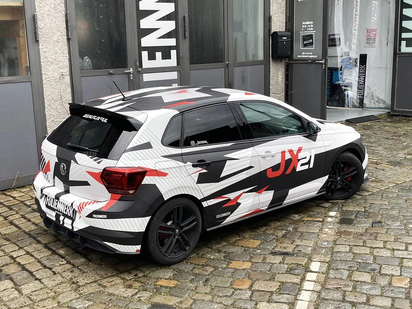 Ein weißes Auto mit schwarzen, roten und grauen Mustern auf gepflastertem Boden vor einer grauen Gebäudetür.