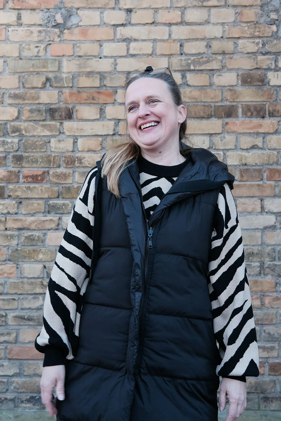 Eine lachende Frau mit langen, braunen Haaren vor einer Ziegelmauer, trägt eine schwarze Weste und ein schwarz-weiß gestreiftes Oberteil.