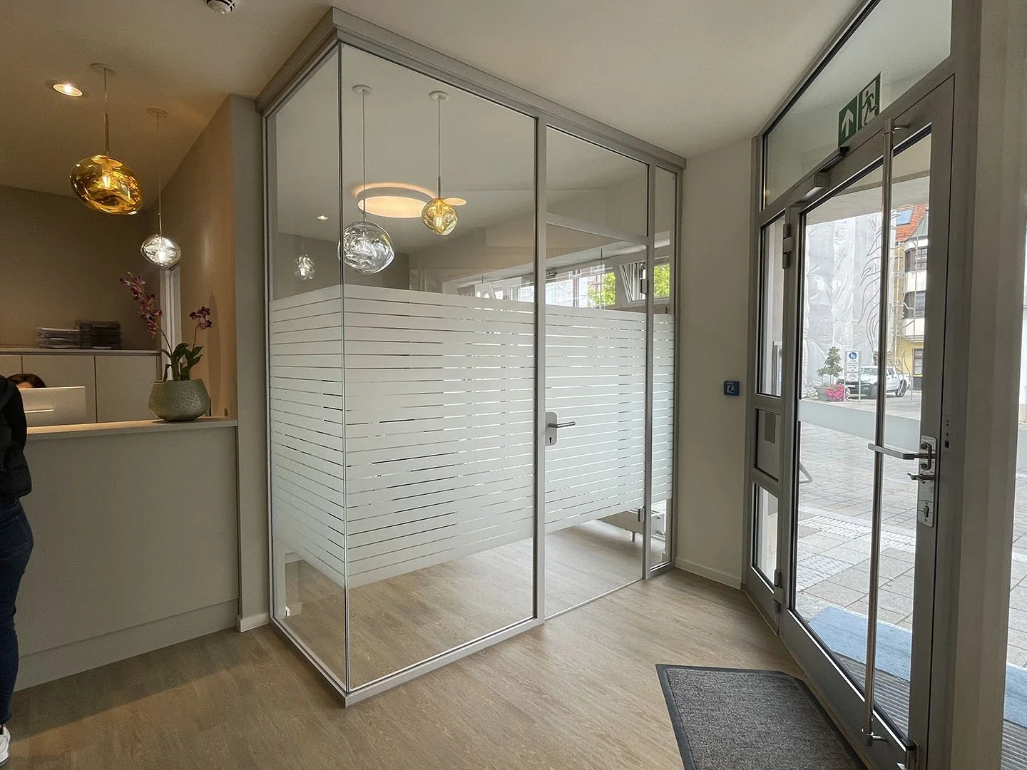 Innenansicht eines modernen Eingangsbereichs mit Glastür und Glasraum mit weißen Holzlamellen. Draußen sind Gebäude, Straße und Fahrzeuge sichtbar.