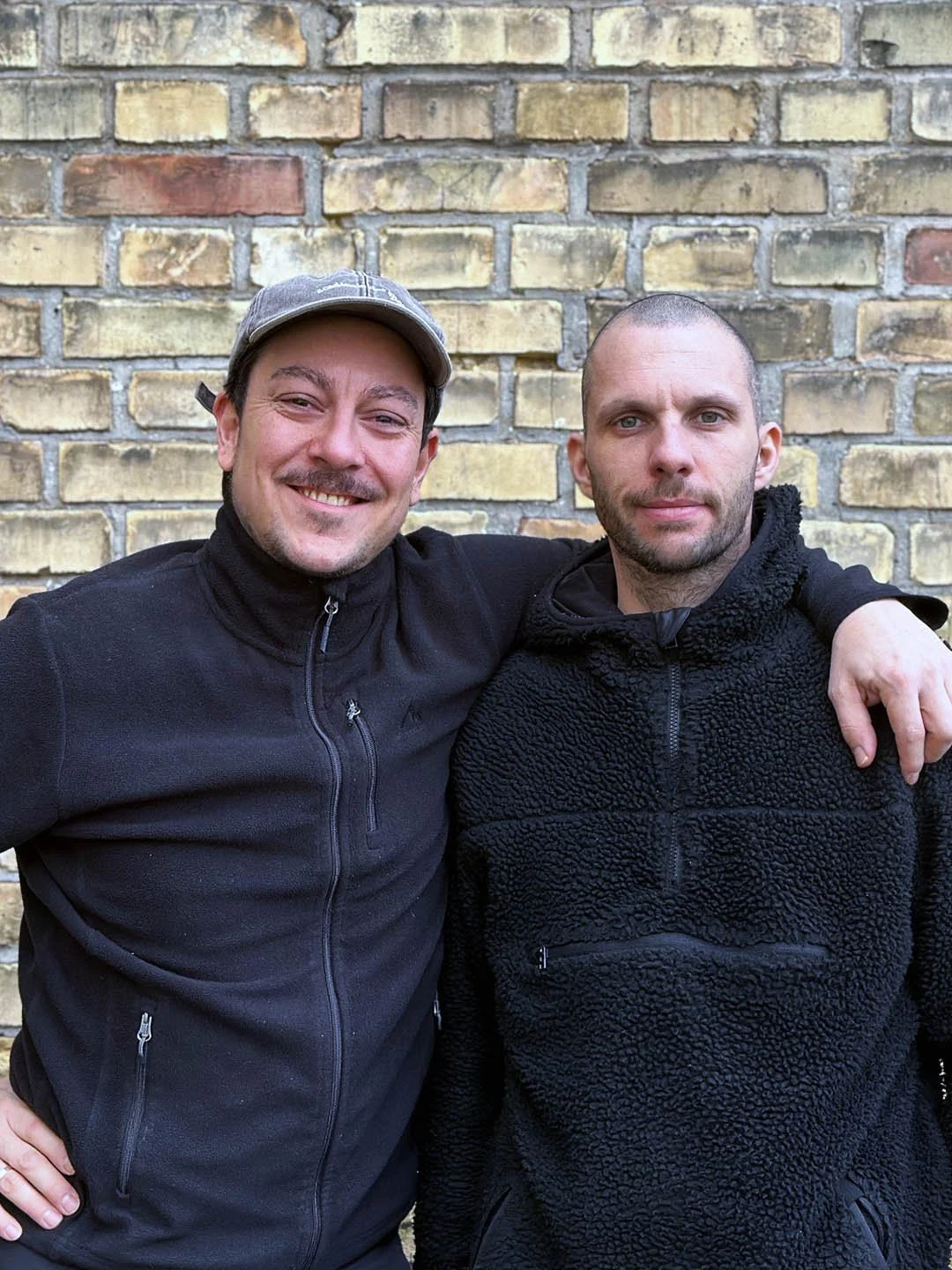 Zwei Männer stehen nebeneinander vor einer Ziegelwand, einer lächelt, der andere schaut ernst, beide tragen schwarze Jacken.