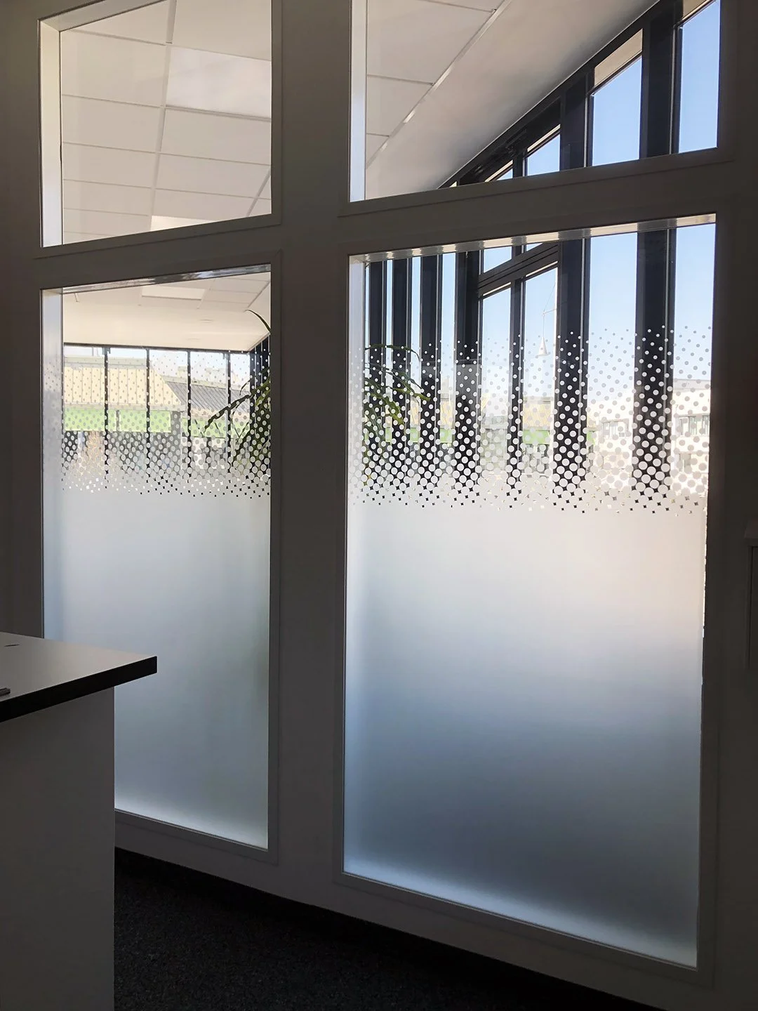 Bürofenster mit Milchglas und einem Muster aus Punkten, blickt nach draußen auf Häuser und Himmel, innen mit dunklem Teppichboden.