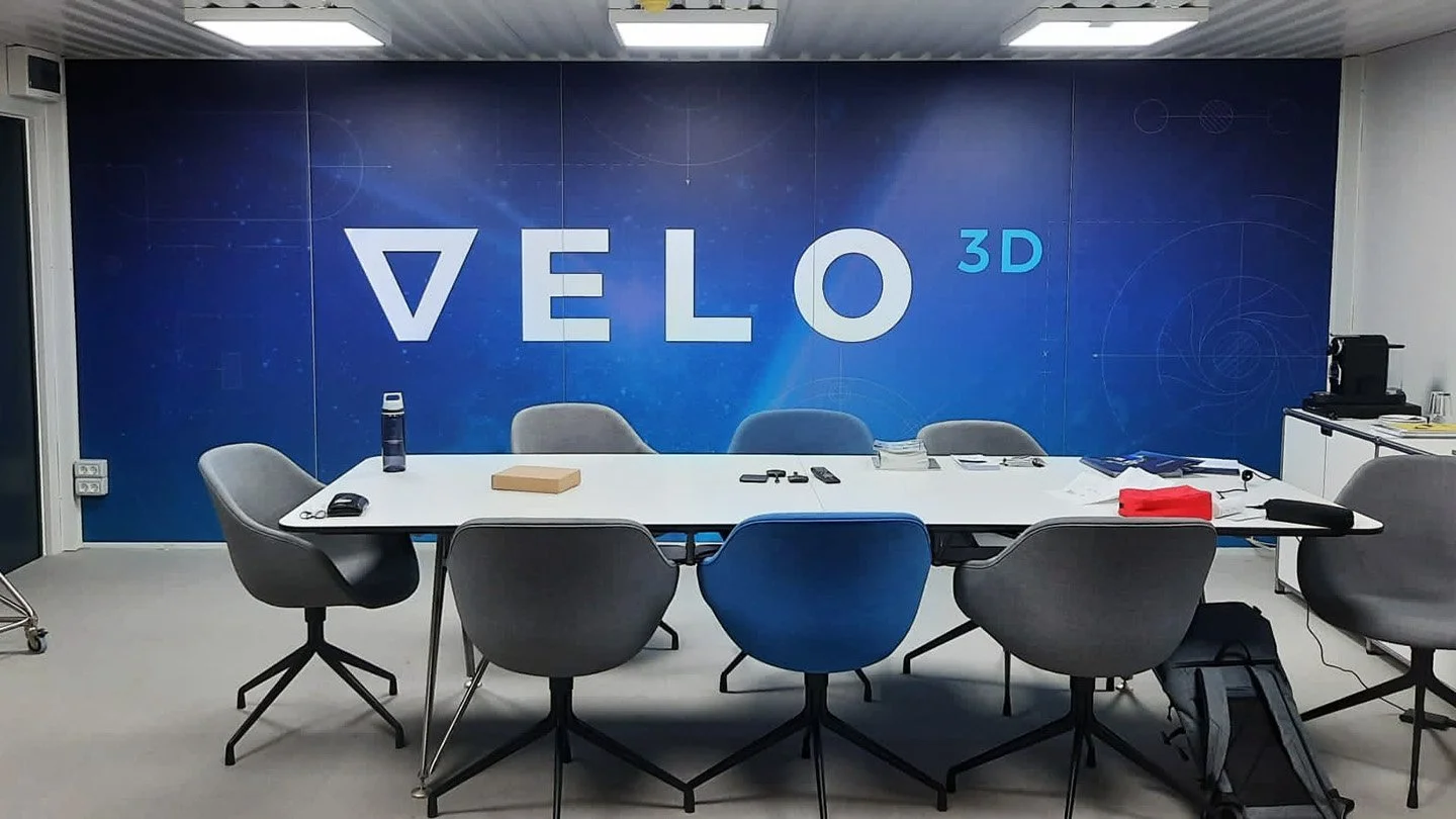 Leerer Konferenzraum mit weißen Tisch, mehreren Stühlen, Wasserflasche, Büchern, Handy und Dokumenten, im Hintergrund eine blaue Wand mit dem Schriftzug "VELO".