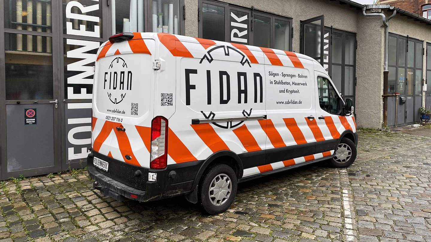 Ein weißer Lieferwagen mit orange-weißen Warnstreifen und Schriftzug "FIDAN" steht auf einem gepflasterten Platz vor einem Gebäude mit großen Fenstern.