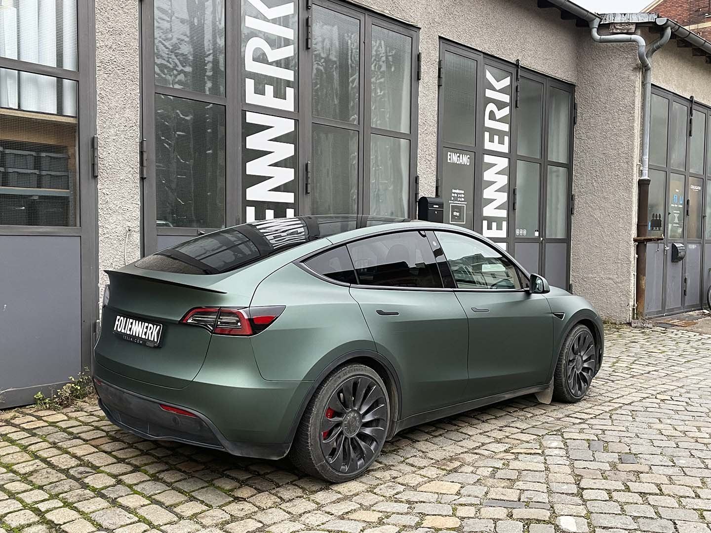 Ein grüner Tesla ist vor einem Gebäude mit Schildern, auf gepflastertem Boden geparkt.