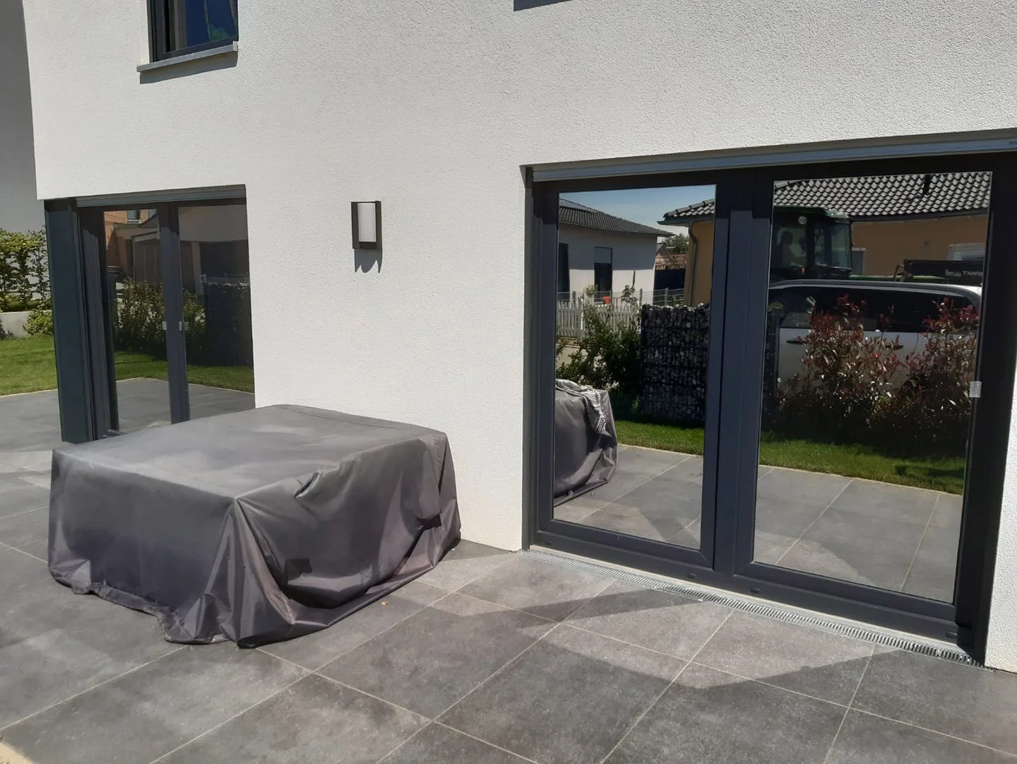 Moderne Terrasse mit grau gefliesten Boden, zwei geschlossene Gartenmöbel unter grauen Abdeckungen, große Glastüren und weiße Hauswand. Im Hintergrund grüne Rasenfläche und Nachbarhäuser.