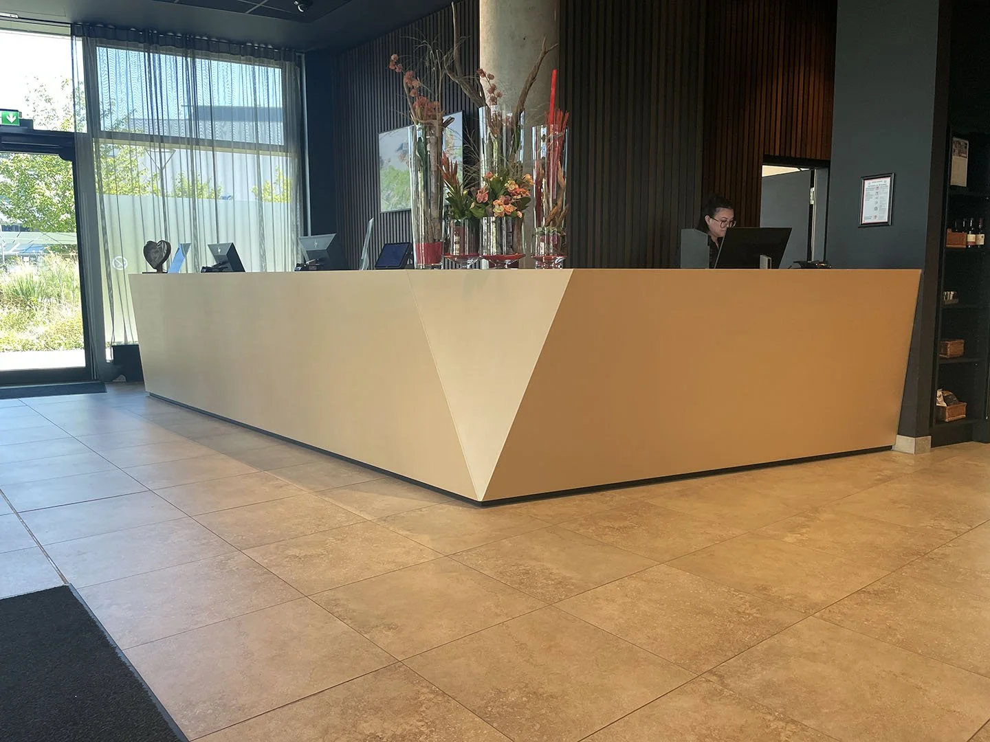 Rezeption in einem Hotel mit gelbem und beigem Front, Dekoration mit mehreren Vasenkombinationen aus Blumen und Zweigen, Mitarbeiterin im Hintergrund am Computer, große Fenster im Hintergrund mit Sicht auf grünen Garten.