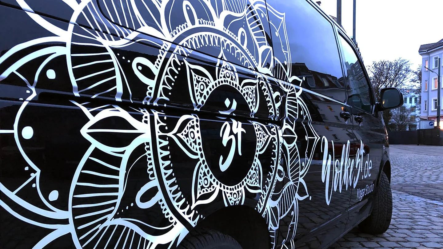 Ein schwarzes Auto mit einer weißen Mandala-ähnlichen Verzierung auf der Seite, die eine symbolische Silhouette enthält, die einer spirituellen Figur ähnelt, neben einigen deutschen Schriftzügen.