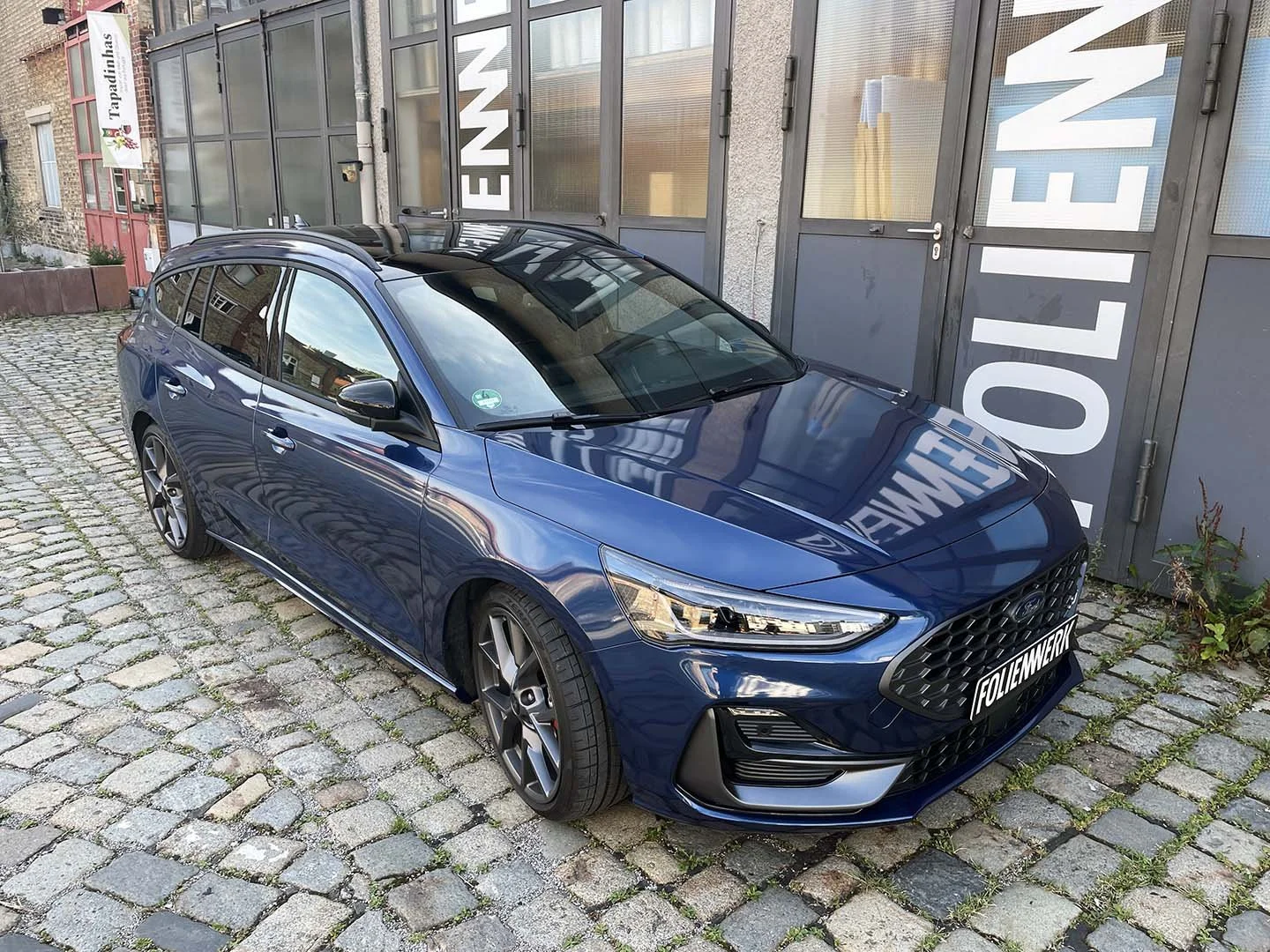 Ein blaues steht auf einer gepflasterten Straße vor einer grauen Tür mit großem Schriftzug, der teilweise in die Kamera spiegelt.