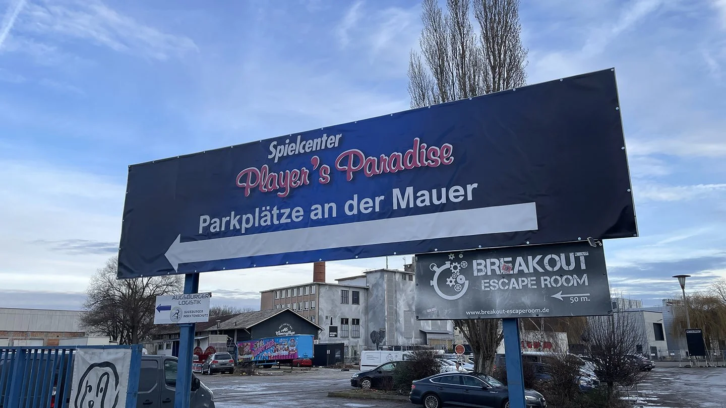 Ein Parkplatz im Freien mit mehreren Autos und einer großen Werbetafel, die auf einen Spielcenter namens 'Player's Paradise' und ein Escape Room hinweist.