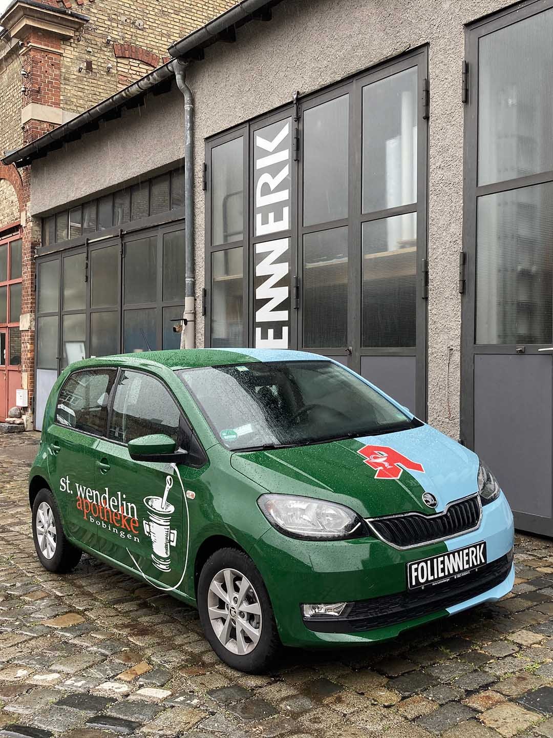Ein grünes Auto mit Werbung für Rotkreuz-Notfalldienst und Apotheke, parkend vor einem Gebäude mit großen Fenstern und einem Schild.