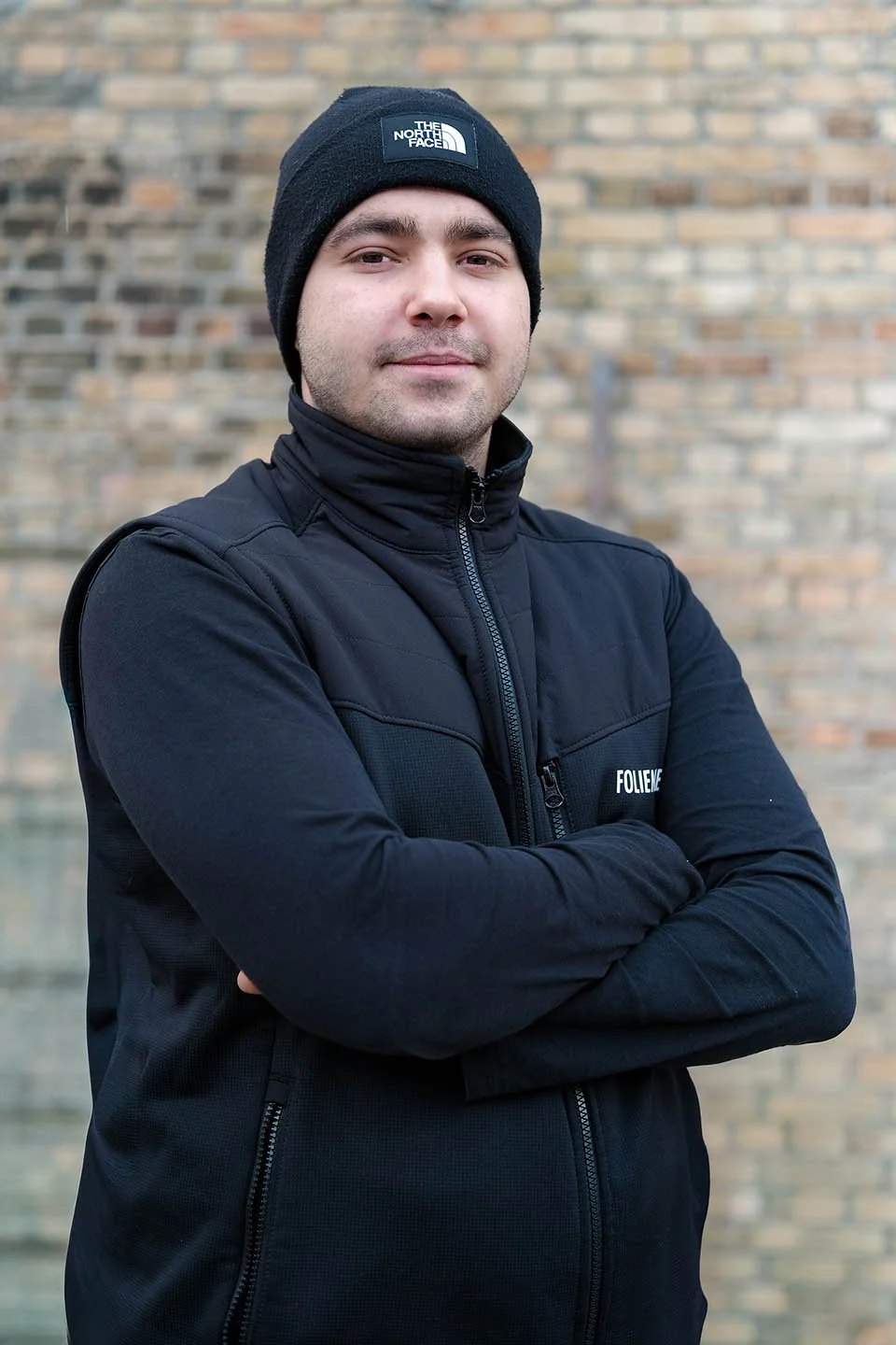Ein junger Mann trägt eine schwarze Jacke und eine schwarze Mütze mit dem Logo von The North Face, steht vor einer gemauerten Wand, mit verschränkten Armen und einem leichten Lächeln auf dem Gesicht.