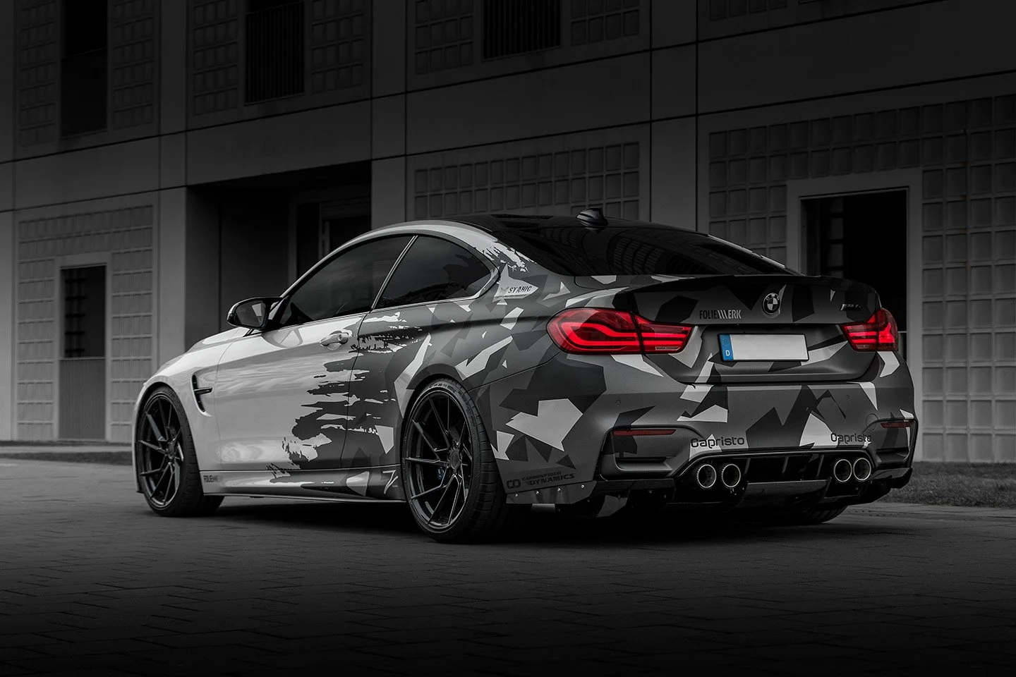 Schwarzer BMW 4er mit Camouflage-Folierung, geteilten Scheinwerfern und Vierrohr-Auspuffanlage, vor einer modernen Gebäudefront.