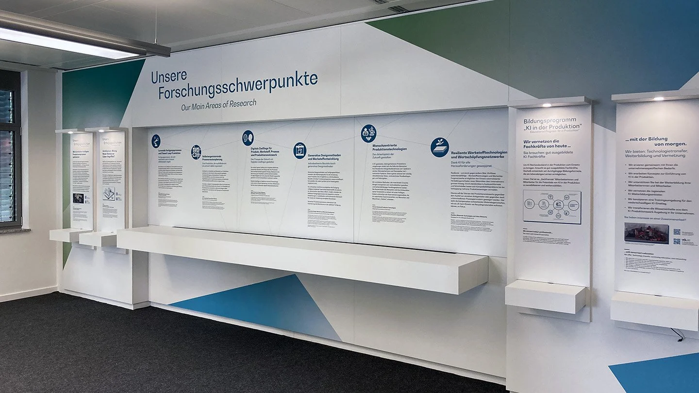 Informationswand mit Texten zu Forschungsschwerpunkten in Deutsch und Englisch, inklusive Diagrammen und Symbolen, in einem Bürogebäude.