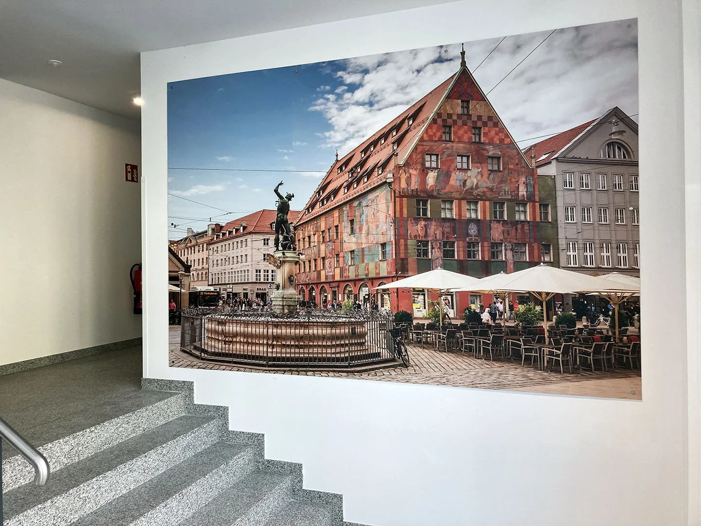Poster mit Brunnen und Statue, umgeben von historischen Gebäuden mit bunten Fassaden, Tischen im Freien und Menschen, die umhergehen. Es ist ein sonniger Tag.
