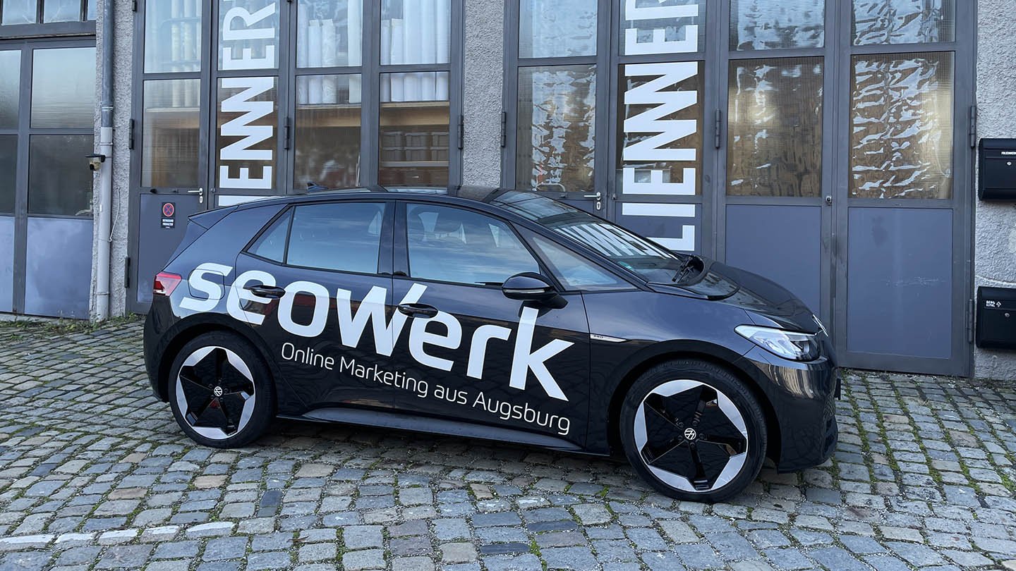 Ein schwarzes Auto mit der Aufschrift "SEOWerK" und "Online Marketing aus Augsburg" ist vor einem Gebäude mit großen Fenstern und einem grauen Türrahmen abgestellt. Das Fahrzeug steht auf einem gepflasterten Bürgersteig.