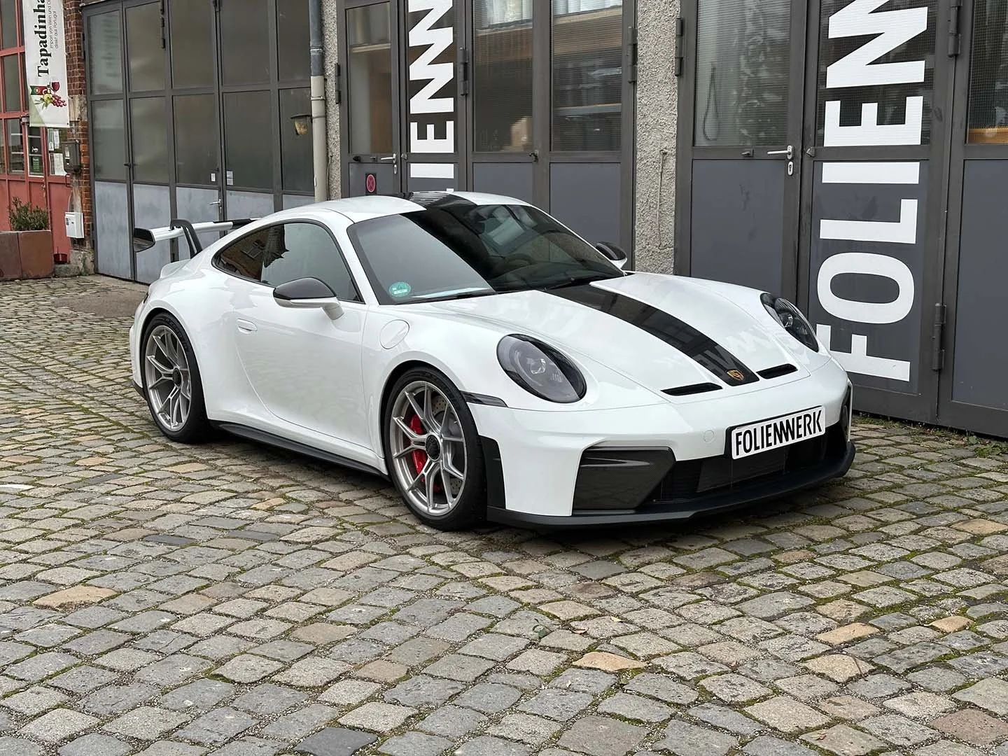 Weiße Sportwagen mit schwarzem Streifen auf der Motorhaube, geparkt auf gepflastertem Boden vor einer Garage mit großen Türen, Beschriftung mit 'FOLIENWERK'.