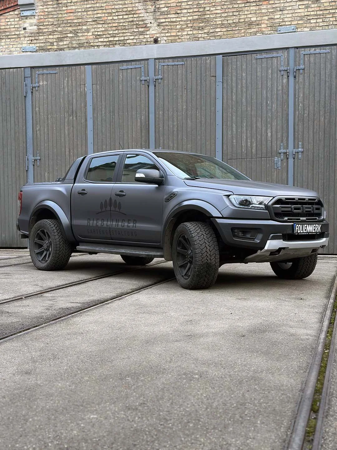 Ein graues Fahrzeug mit Firmenlogo und Schriftzug auf der Seite, vor einer hohen Holztür, auf einem gepflasterten Boden mit Eisenbahnschienen.