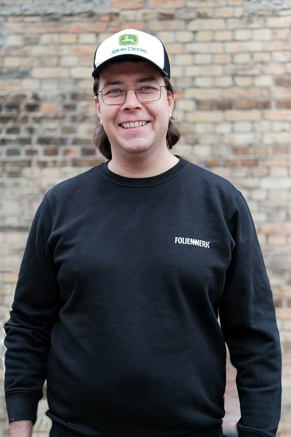 Lächelnder Mann mit Brille, schwarzem Sweatshirt mit Aufdruck 'FOLIENWERK' und einer weißen Kappe mit dem John Deere Logo, vor einer Backsteinmauer.