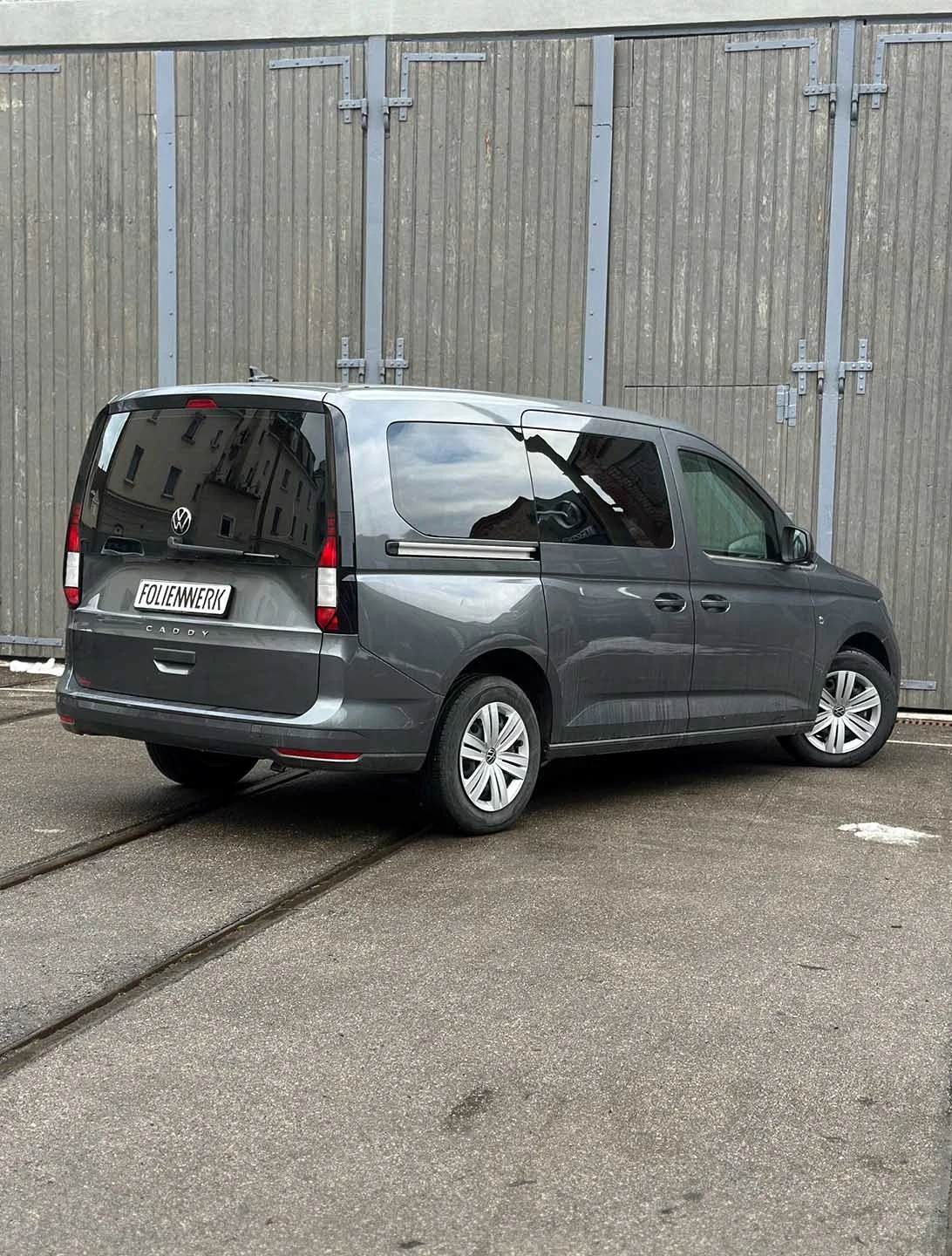 Ein grauer Volkswagen Caddy wird in einem Parkplatz mit Schienen für eine Straßenbahn oder einen Transportschlitten geparkt, vor einer Holz- und Metallwand.