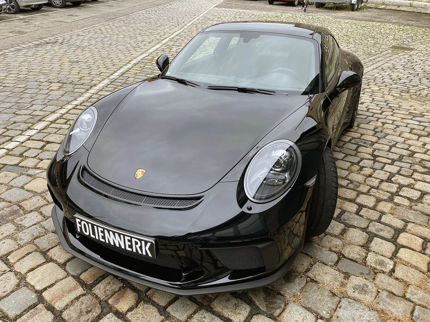 Schwarzer Porsche-Sportwagen mit Kennzeichen "FOLIENWERK" auf gepflastertem Parkplatz.