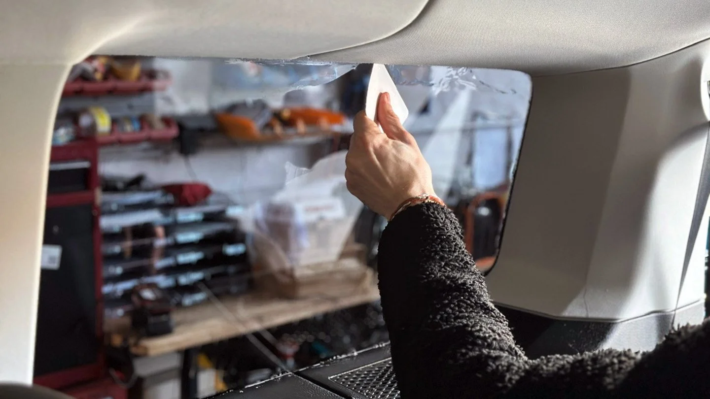 Eine Hand wischt mit einem Papiertuch gegen das Fenster eines Flugzeugs, während im Hintergrund Werkzeuge und Werkbänke sichtbar sind.