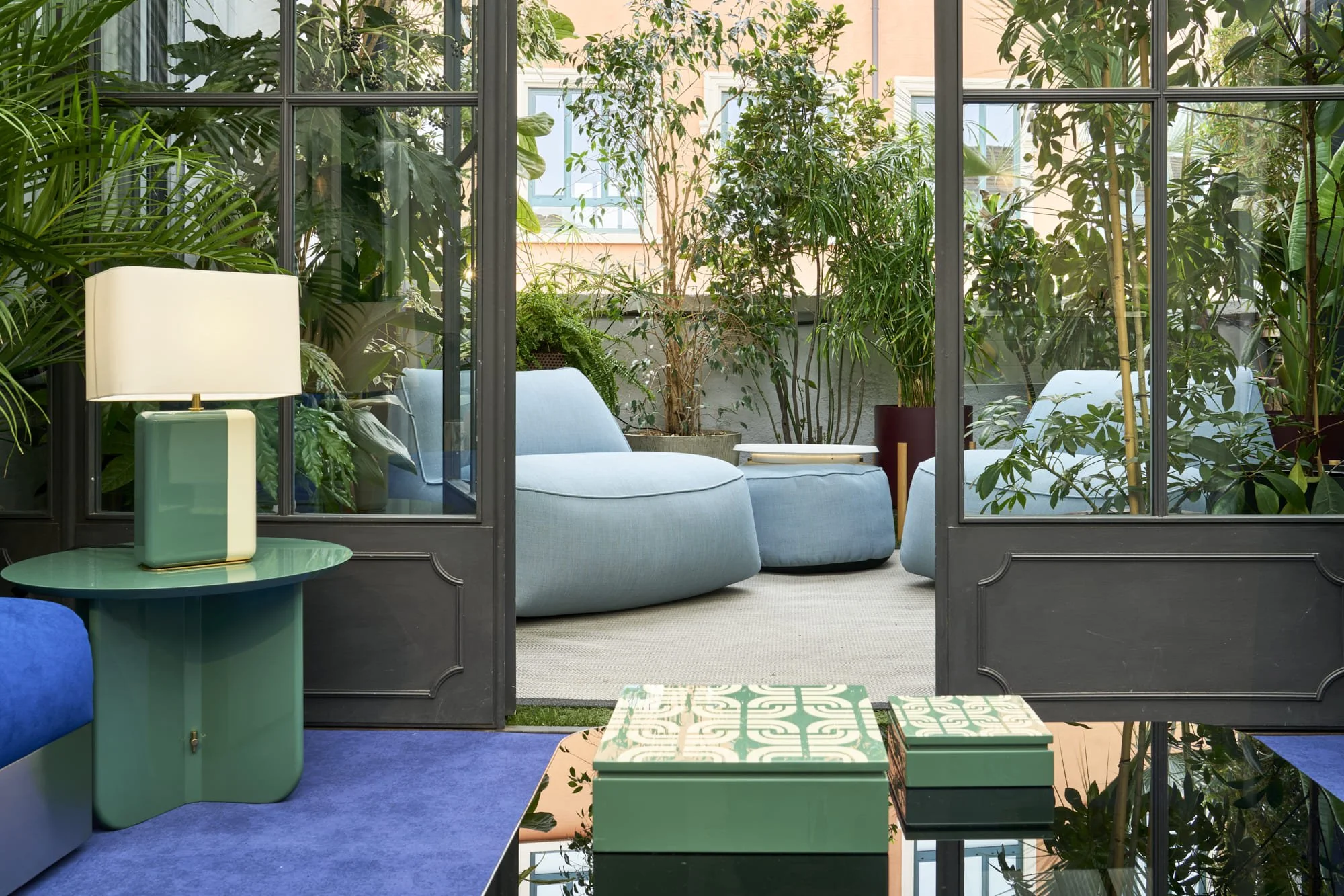 Stylish indoor sitting area with light blue chairs and green plants, separated by glass doors from a patio with more greenery and outdoor furniture.