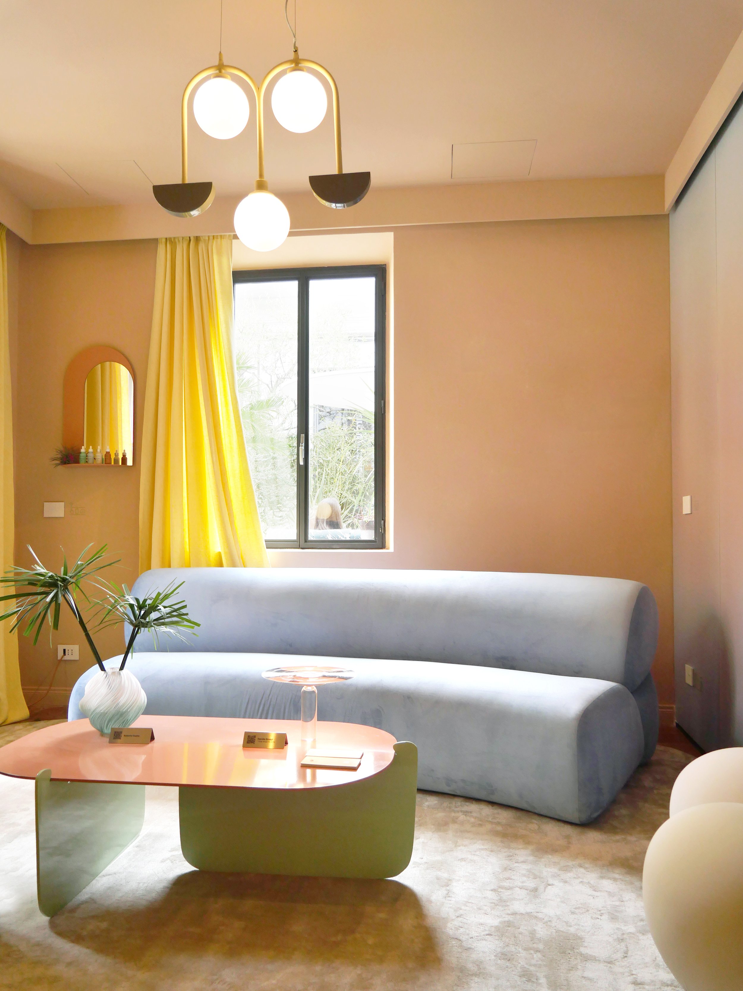 Modern living room with a beige wall, a large window with yellow curtains, a light blue sofa, a pink coffee table with two gold name plaques, a green chair, a potted plant, a small mirror on the wall, and a contemporary chandelier with white globes.