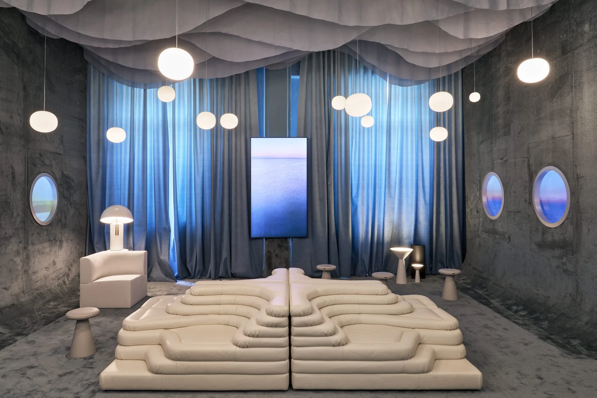 Modern interior room with gray textured walls, blue curtains, and a ceiling decorated with hanging white spherical lights. There is a large vertical digital display screen on the back wall showing a serene landscape. The furniture includes a white, layered, stepped sofa in the center, a white chair with a side table and lamp on the left, and three small, tiered side tables on the right. The room has two porthole windows and a ceiling with cloud-like white shapes.