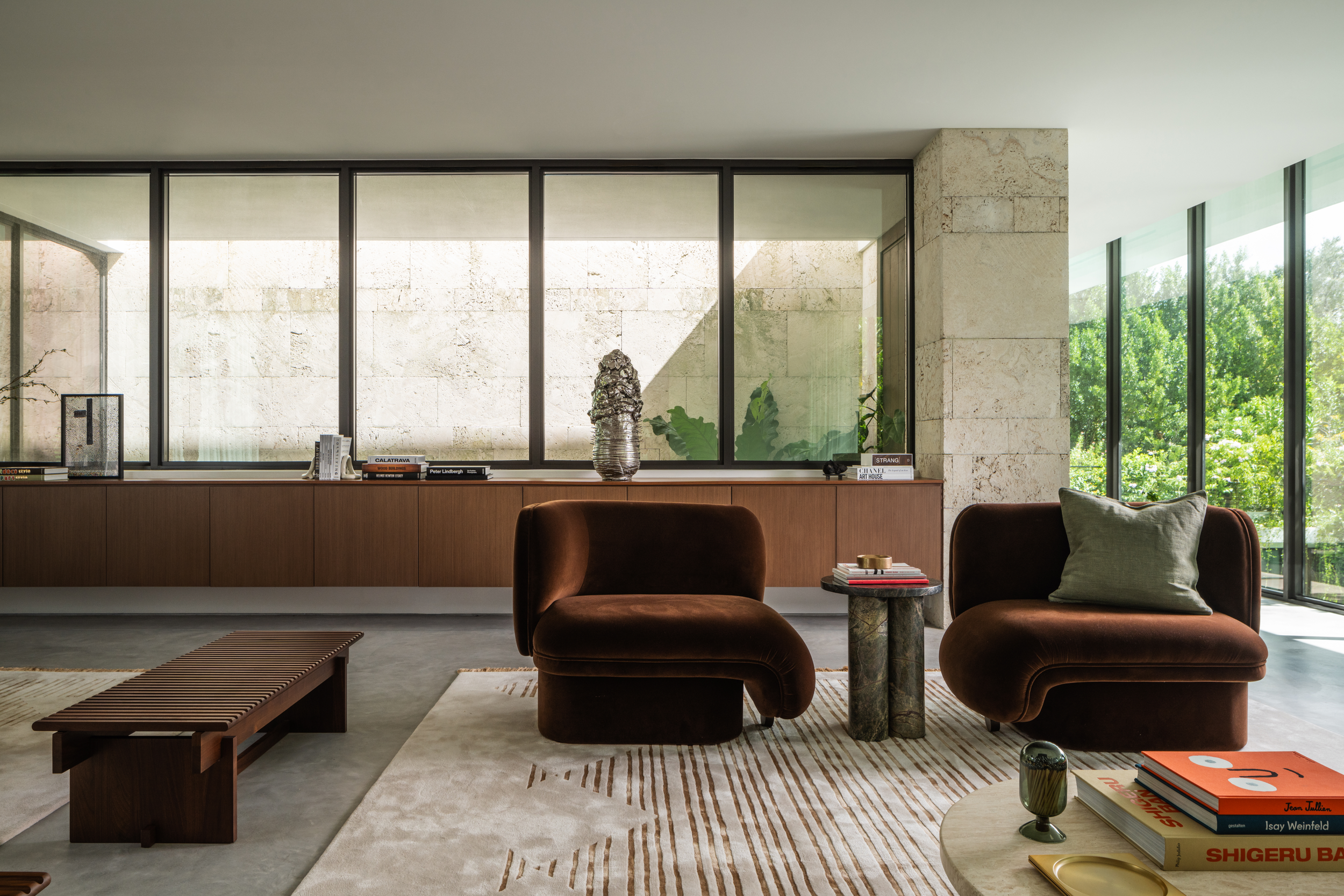 Modern living room with large windows, two brown velvet chairs, a stone side table with books and decor, a striped rug, and a wooden bench, with greenery visible outside.