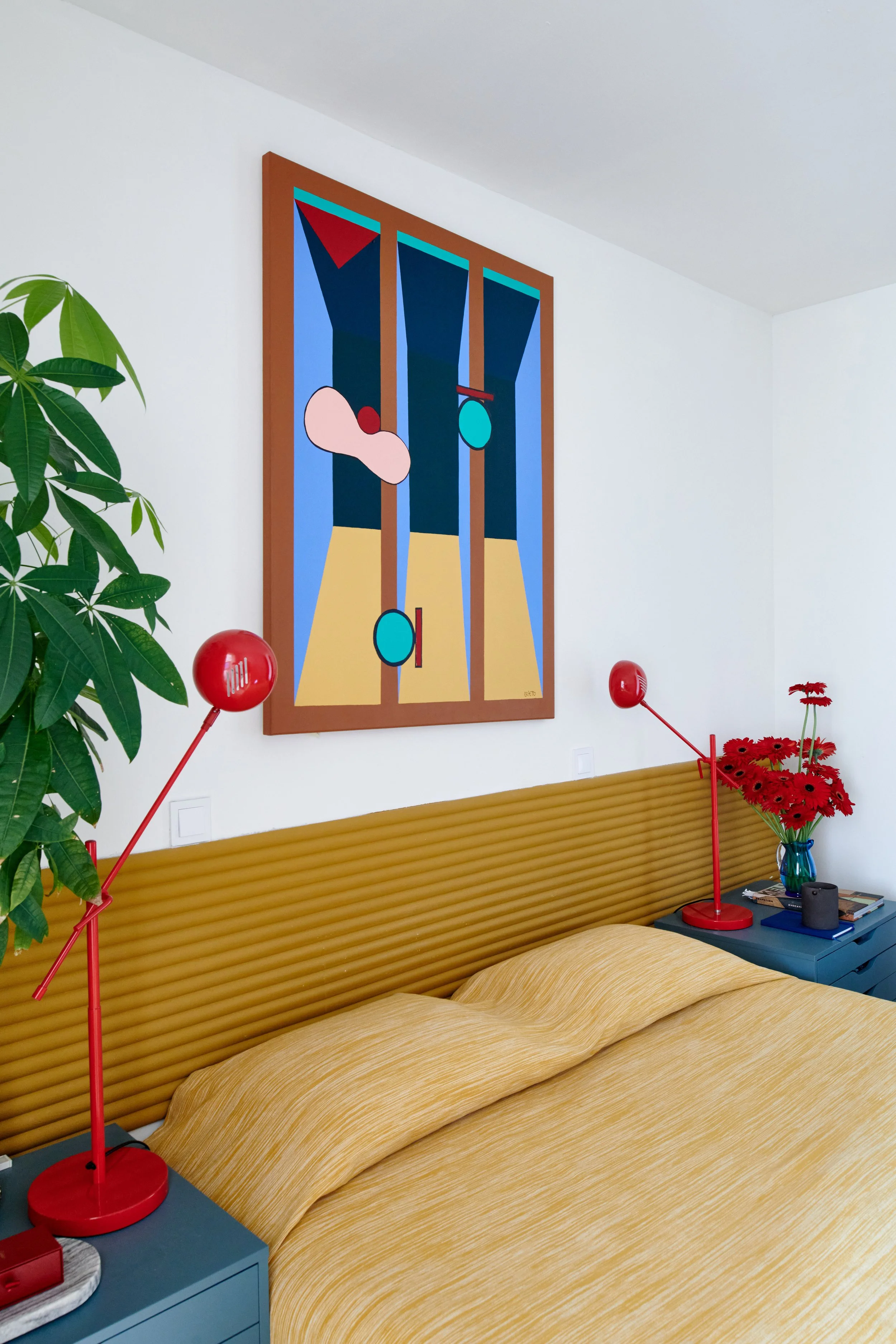 Interior of a bedroom with a mustard yellow bedspread, modern yellow headboard, two red bedside lamps, a blue bedside table with books, a red vase with red flowers, a large green houseplant, and a large colorful abstract painting on white walls.