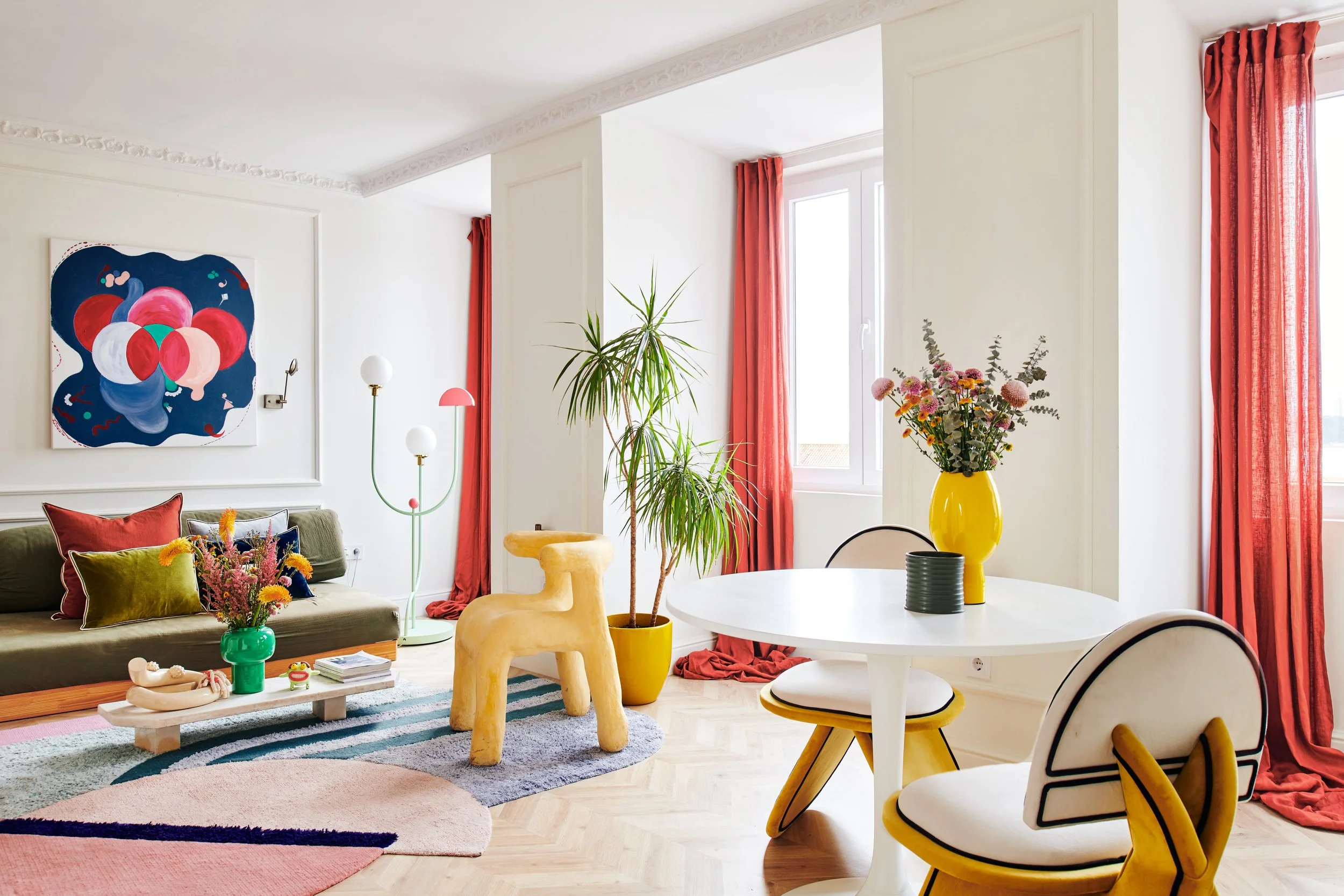 Bright, modern living room with white walls, large windows with red curtains, a green sofa with colorful pillows, a colorful abstract painting, a multicolored floor lamp, a round white dining table with two yellow-accented chairs, a yellow vase with flowers, a large potted plant, and a mix of pastel and multicolored rugs.