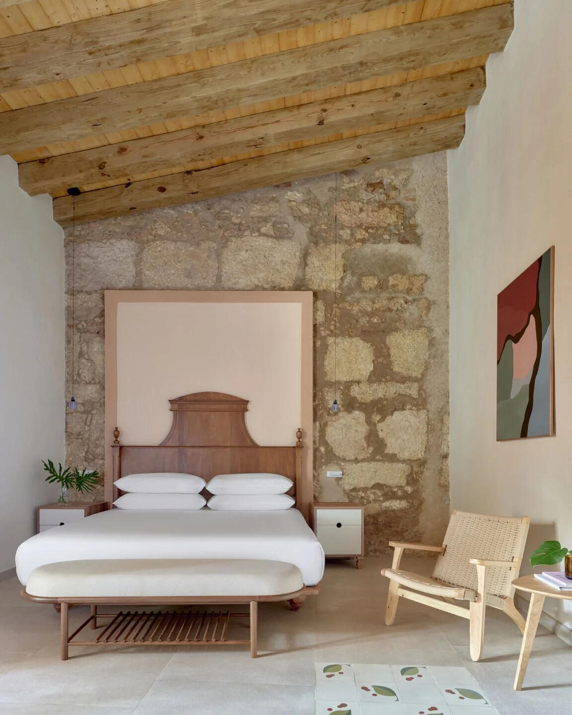 A bedroom with a wooden bed, white bedding, and four pillows, flanked by nightstands, one with a plant. A beige armchair and a small round table with a plant and books are near the wall. The room has a stone wall and exposed wooden ceiling.