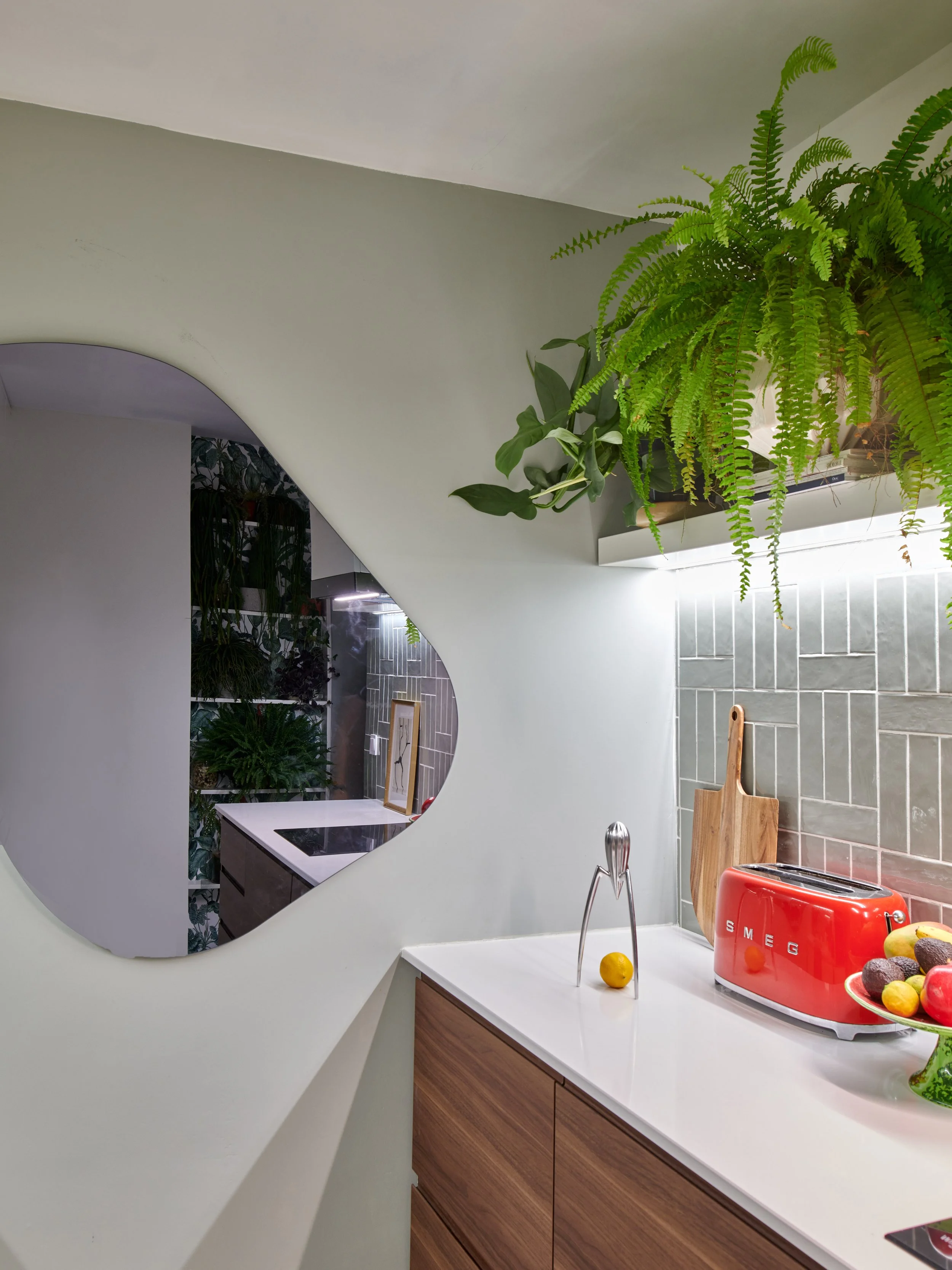 Kitchen countertop with red toaster, wooden cutting board, decorative fruit bowl, and hanging green plants above mirror and backsplash.