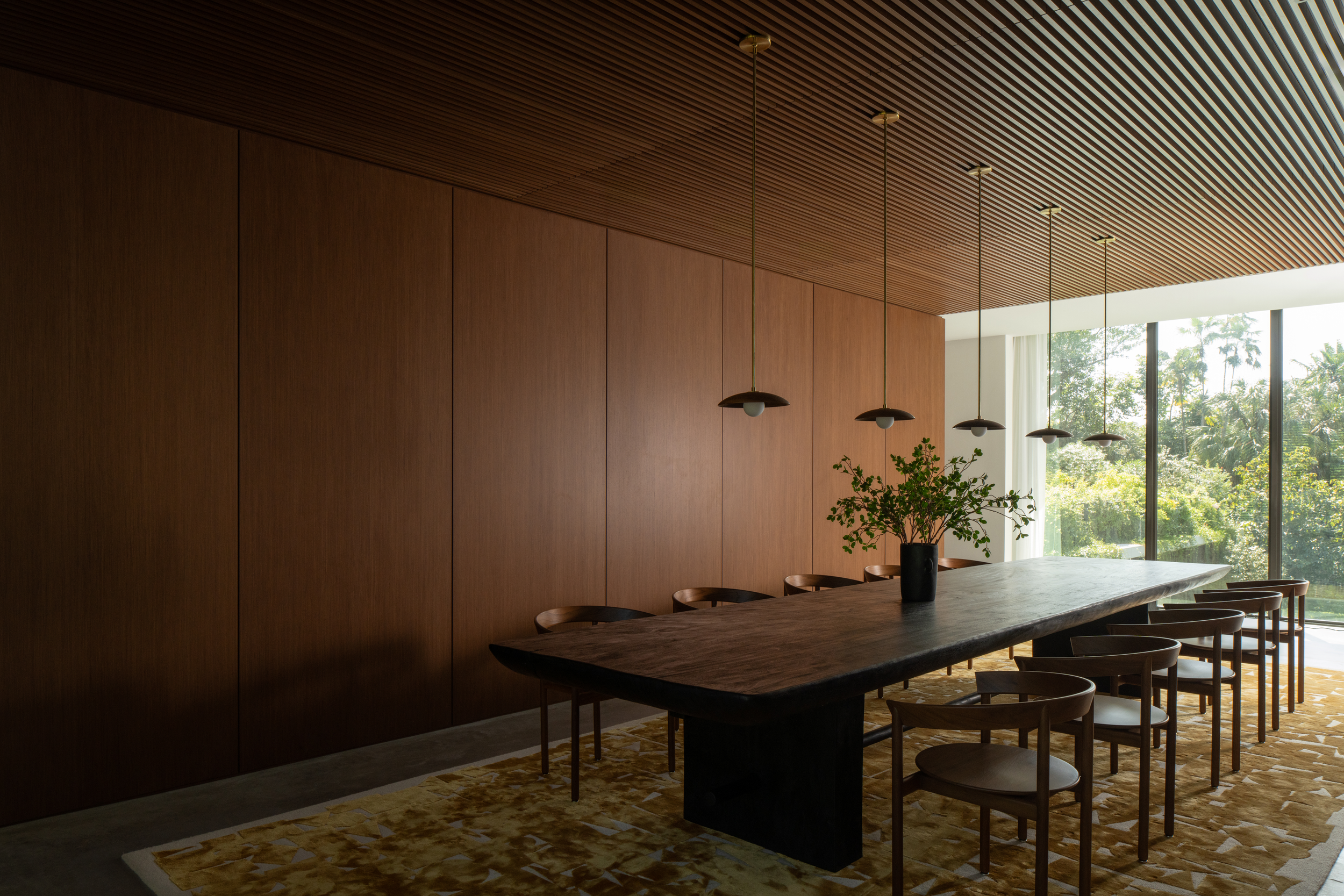 Modern dining room with a large wooden table, chairs, hanging pendant lights, a black vase with green plants, a large window with trees outside, and a patterned rug.